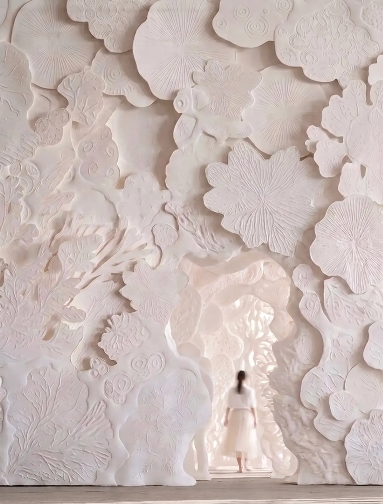A second visualisation of the same white relief wall, framed wider. The overlapping discs and fronds fill the full height, with the arched opening and the figure at its centre.