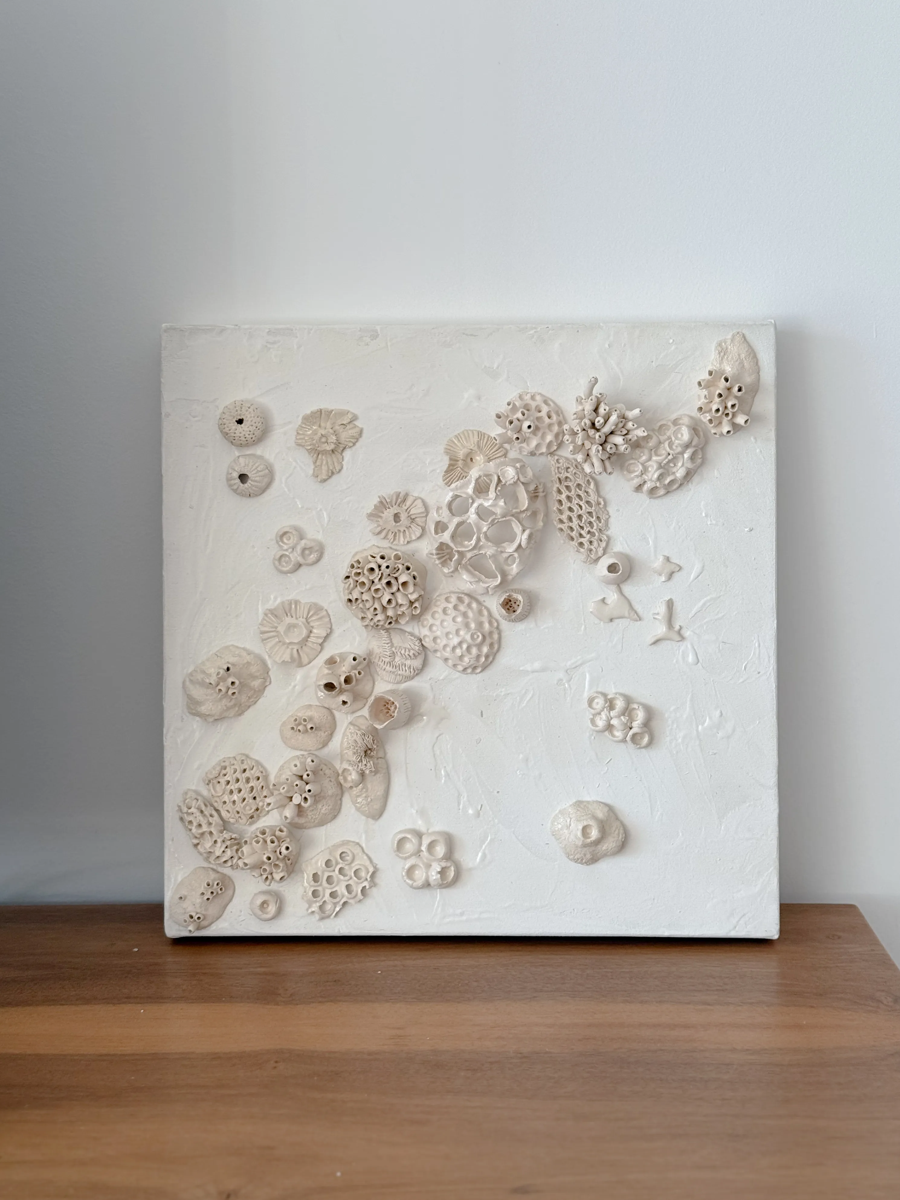 Square canvas resting on a wooden surface against a pale wall. Bone white ceramic rosettes and perforated discs drift diagonally across a textured white ground.