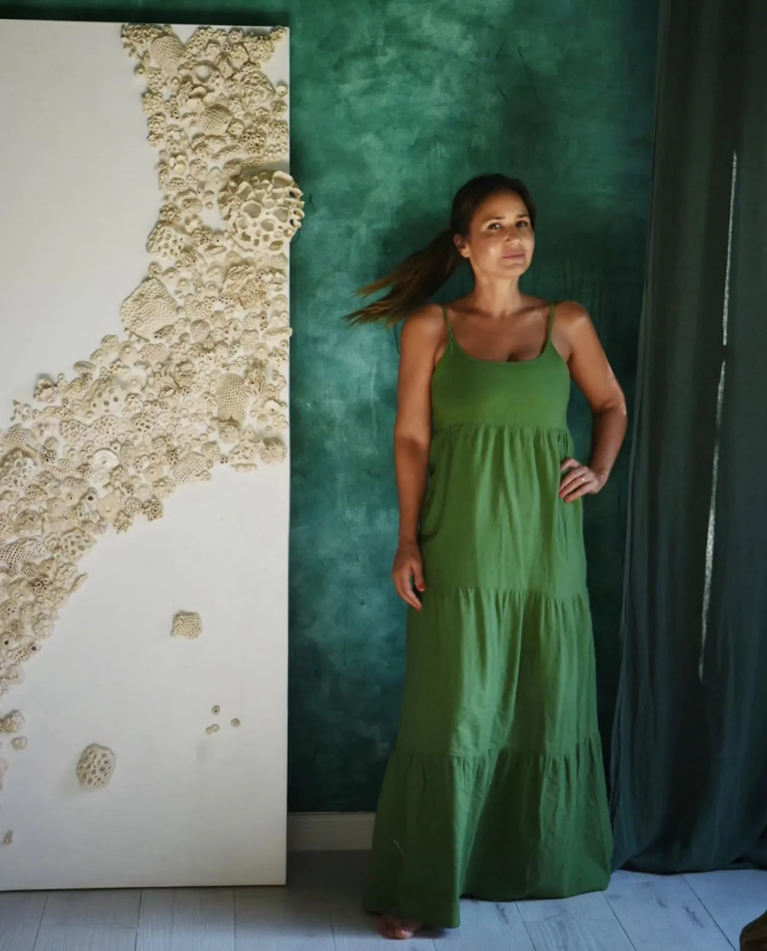 Marina Maiztegui standing beside a tall Corals canvas, a drift of pale ceramic forms across white, against a deep green wall.