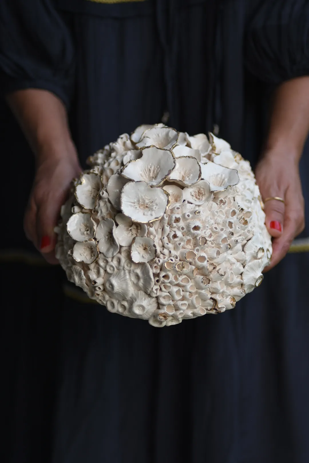 Rounded ceramic reef form held in two hands against a dark grey dress. Cupped discs cluster over a crown of fine open tubes.