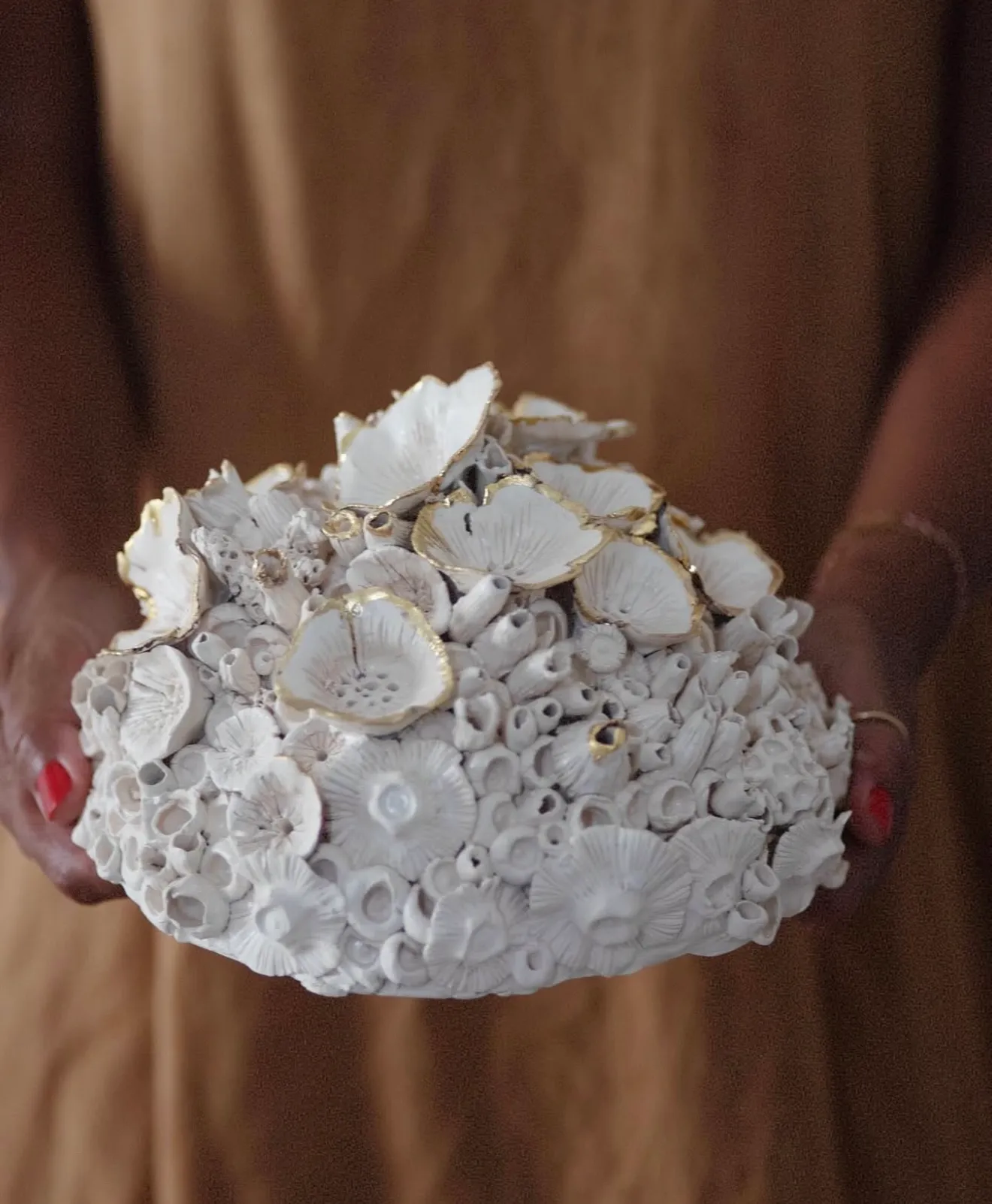 Ceramic reef form held in two hands against an ochre dress. Discs rimmed in gold luster gather at the top over a dense field of pale tubes.