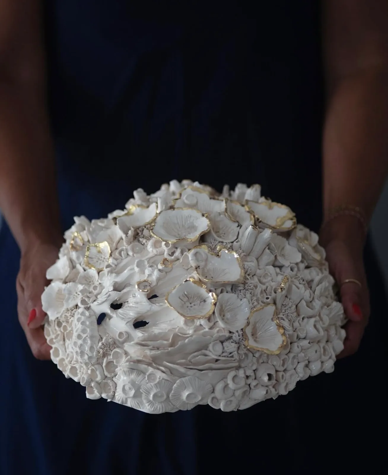 Oval ceramic reef form held in two hands against a navy dress. Gold rimmed cups spread across the crown, a few pierced by small dark openings.