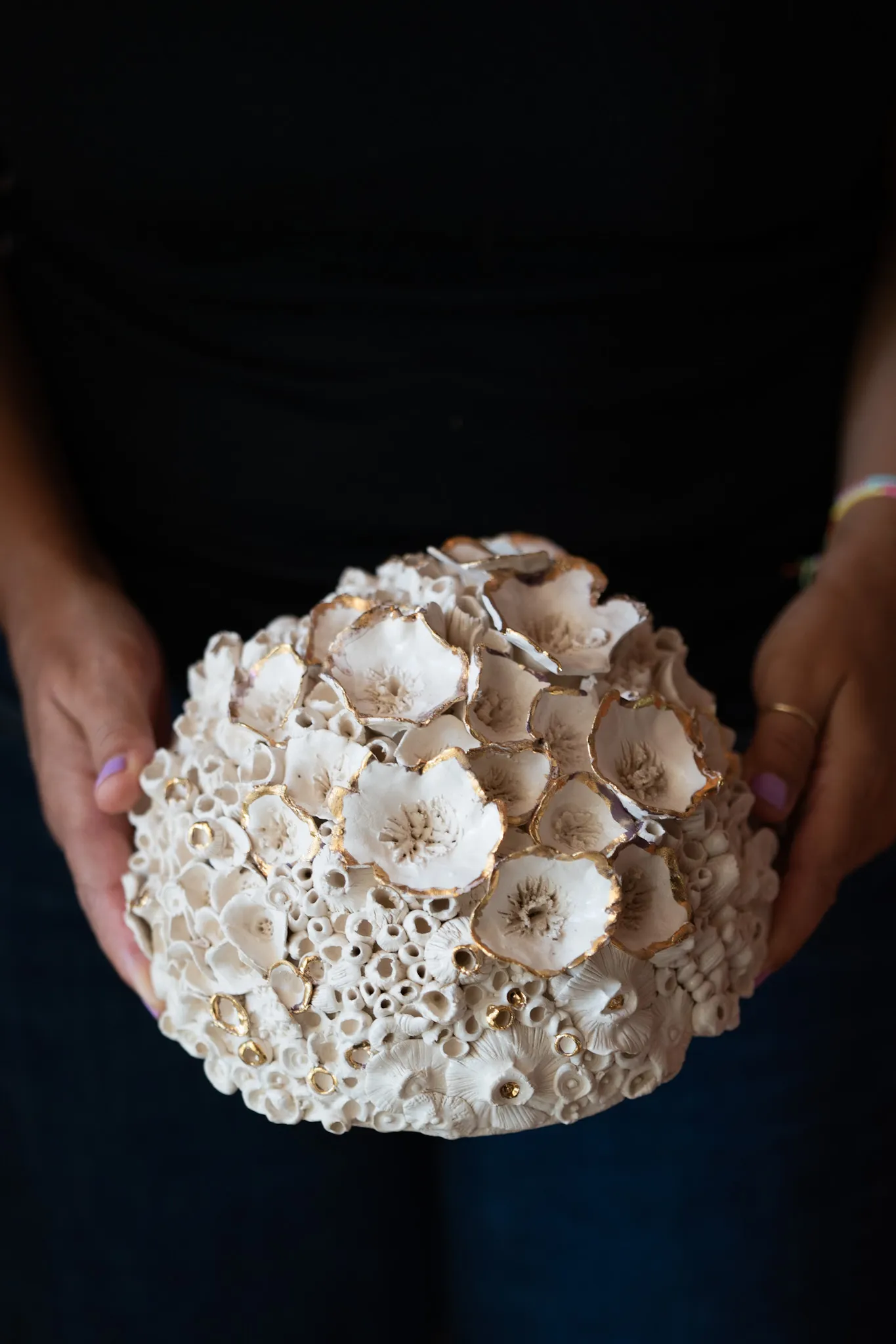 Domed ceramic reef form held in two hands against a black top. Cupped discs and barnacle tubes crust the surface, some rims touched with gold luster.