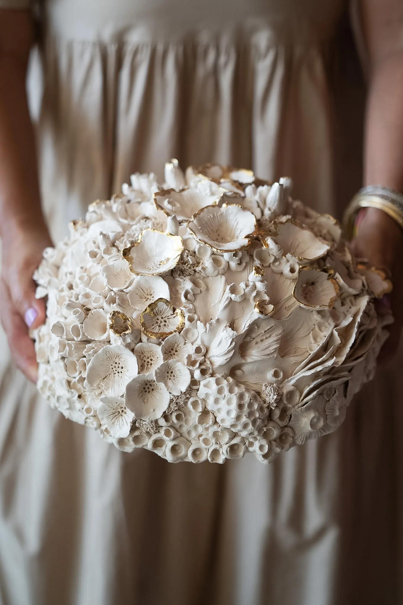 Rounded ceramic reef form held in two hands against a sand coloured dress. Open cups and clustered tubes build up across the surface in cream and bone white.