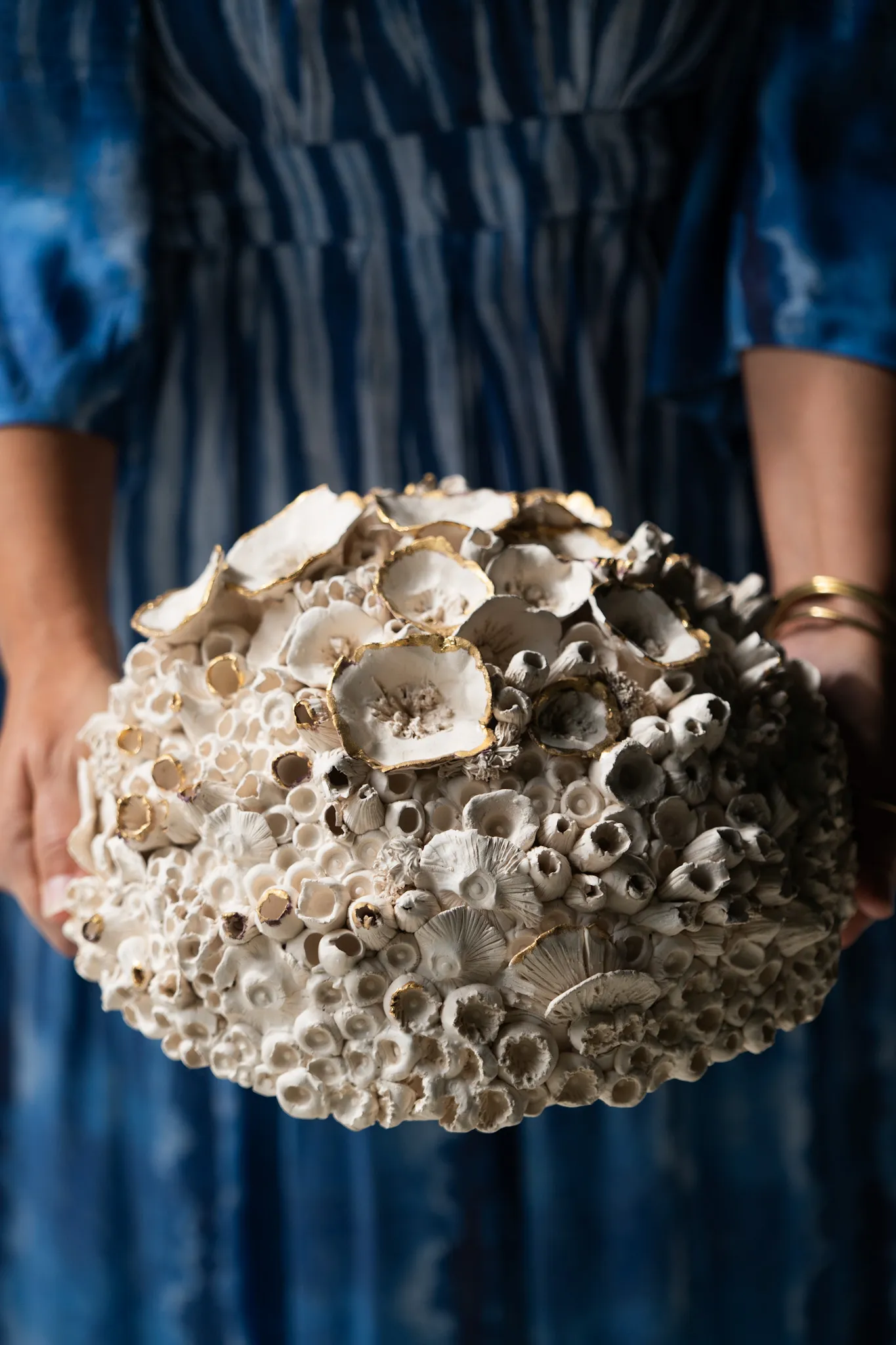 Oval ceramic reef form held in two hands against a blue tie dyed dress. Dense barnacle clusters and cupped discs cover it, flecked with gold luster.