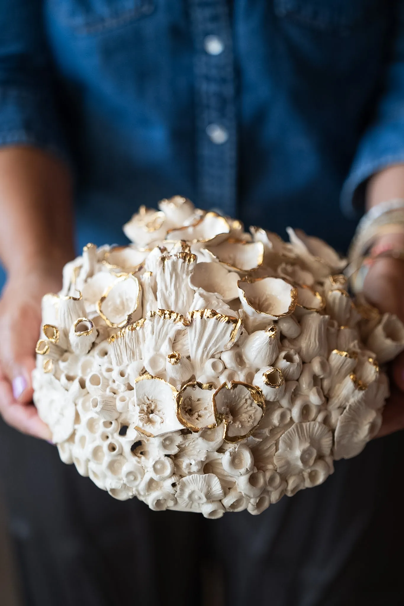 Domed ceramic reef form held in two hands against a denim shirt. Frilled cups and open tubes crust the surface, several rimmed in gold.