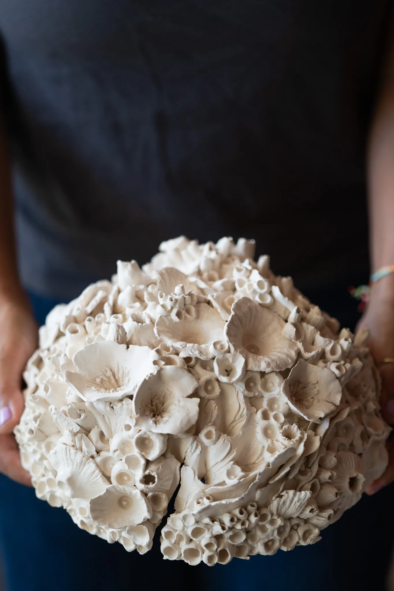 Domed ceramic reef form held in two hands against a grey top. Large open flower like cups sit among fine tubes in matte cream.