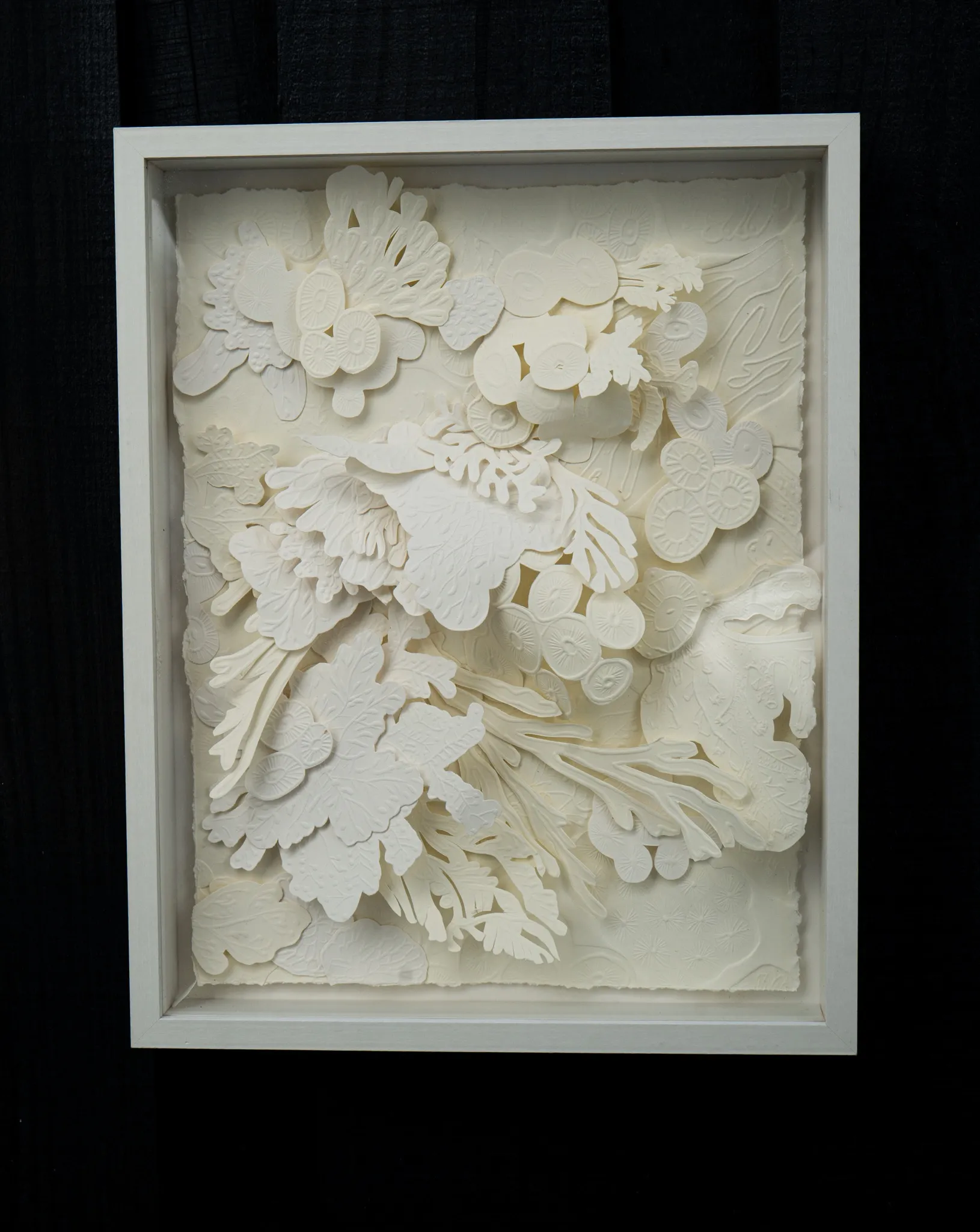 White embossed and hand-cut cotton paper relief in a white box frame on a black wall. Clusters of discs, blossoms and fronds rise in layers across the sheet, the deckled edge of the paper visible inside the frame.