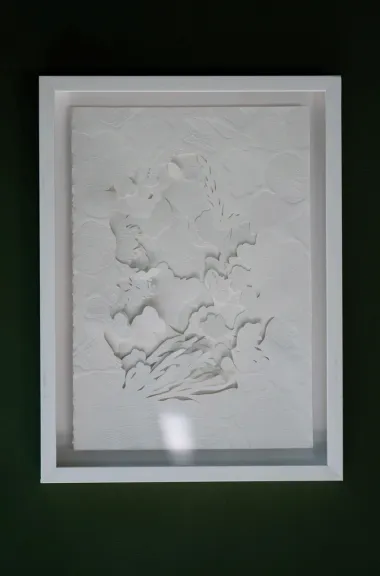 White embossed and hand-cut cotton paper relief in a white box frame on a dark green wall. Layered forms gather at the centre and break into fine ribbon-like cuts along the lower edge.