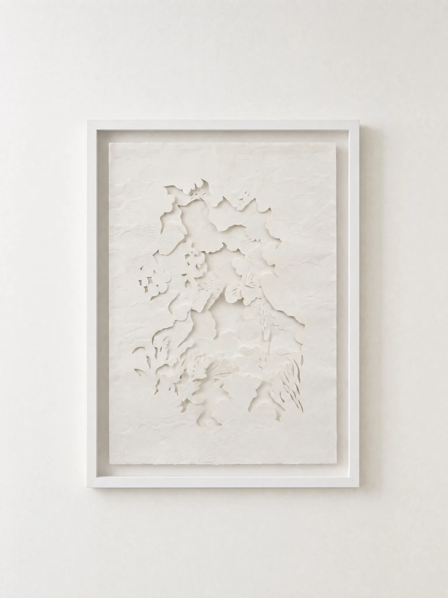 White embossed and hand-cut cotton paper relief in a slim white frame on a white wall. Cut and lifted shapes gather down the centre of the sheet, with a wide margin of plain paper around them.