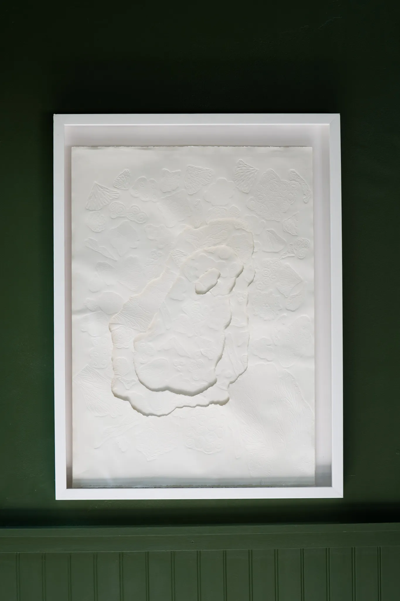 White embossed and hand-cut cotton paper relief in a white box frame on a dark green wall. Soft rounded forms overlap at the centre of the sheet in low relief, ringed by fainter embossed shapes.