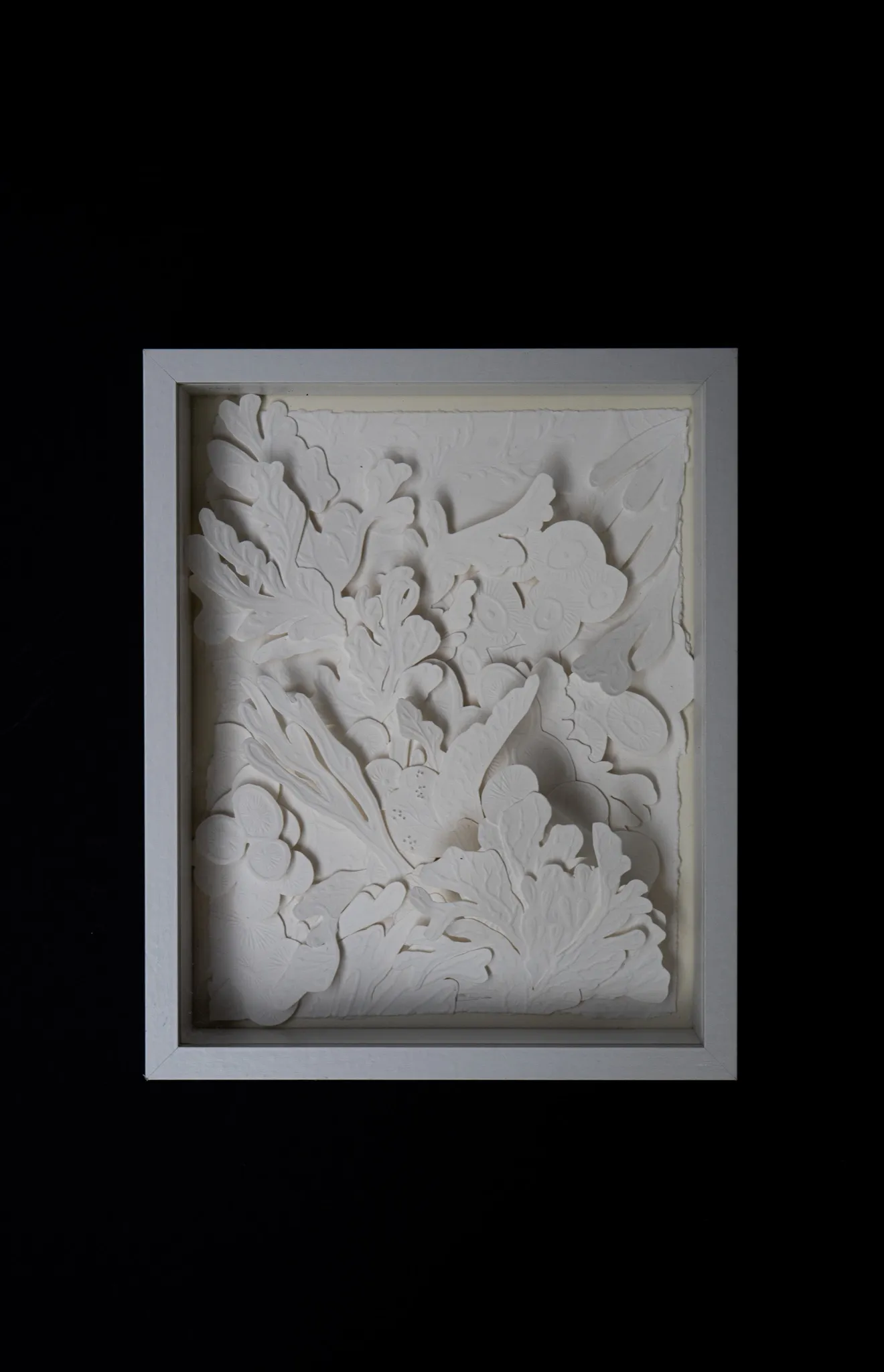 White embossed and hand-cut cotton paper relief in a white box frame on a black background. Lobed leaves, fronds and rounded blossoms crowd the whole sheet in layers.